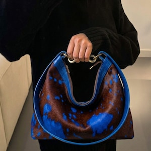 Puede incluir: Un bolso de estilo hobo con un patr&oacute;n de piel de vaca en marr&oacute;n y azul con un borde de cuero azul liso. El bolso tiene un asa circular grande de cuero azul unida a un conector de metal en tono dorado en la parte superior central de la abertura.