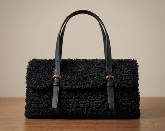 May include: A black handbag crafted from textured, curly faux-shearling material. The bag features a rectangular flap closure secured by two vertical black straps with small, metallic gold-toned buckles and two matching black top handles.