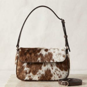 May include: A shoulder bag featuring a brown and white cowhide pattern with a flap closure. The bag has a dark brown leather strap with an adjustable buckle and a matching detachable strap lying flat, both featuring silver-toned metal hardware.