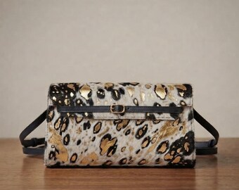 May include: A rectangular crossbody bag featuring a textured animal print pattern in black, white, and metallic gold. The bag includes a thin black leather strap with a small gold-toned metal buckle centred horizontally across the front flap.