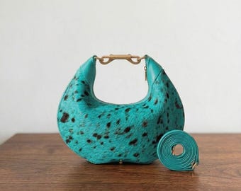 May include: A crescent-shaped handbag featuring a vibrant turquoise dyed hair-on-hide texture with scattered black spots. The bag includes a gold-toned metal top handle with a central clasp mechanism and a matching detachable turquoise shoulder strap.