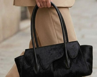 May include: A black handbag with a textured, hair-on-hide finish and a structured silhouette. The bag features two long, thin black leather handles and a distinctive scalloped flap design across the front, complemented by a beige blazer.