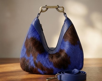 May include: A hobo-style handbag featuring a blue and dark brown cowhide pattern with a smooth blue leather trim. The bag has a unique antique brass metal top handle with a clasp mechanism and includes a detachable blue leather shoulder strap.