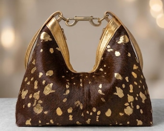 May include: A hobo-style handbag crafted from dark brown hair-on hide featuring an irregular gold metallic splatter pattern. The bag includes gold leather trim along the top edges and a central gold-toned metal lobster clasp closure hardware.