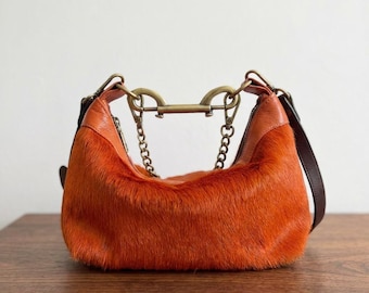 May include: A vibrant orange hobo bag crafted from textured hair-on hide leather. The bag features smooth orange leather accents, a dark brown shoulder strap, and a distinctive antique brass-toned metal hardware connector with two chains.