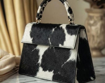 May include: A structured handbag featuring a black and white cow print pattern with a textured hair-on-hide finish. The bag has a single top handle, a fold-over flap closure, and gold-toned hardware attachments for a detachable shoulder strap.