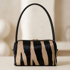 May include: A cylindrical handbag featuring a black and beige zebra-striped pattern with a textured hair-on-hide finish. The bag includes a single black arched handle and a top zipper closure with gold-toned hardware, presenting a structured shape.