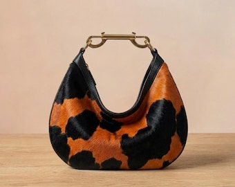 Puede incluir: Un bolso de mano en forma de media luna con un patrón de piel de vaca texturizada con manchas negras y naranjas. El bolso incluye un borde de cuero negro liso en la parte superior y un asa rígida de metal dorado unida al marco superior.