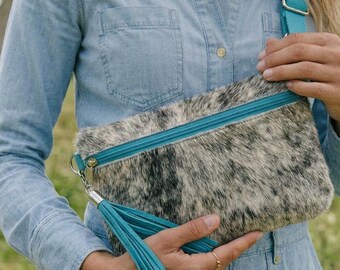 May include: A rectangular crossbody bag featuring a speckled black, white, and grey cowhide texture. The bag includes a bright turquoise zip closure and a matching turquoise leather tassel attached to the zip pull, with an adjustable strap.