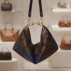 Könnte beinhalten: Eine Hobo-Handtasche mit einer braun-blauen Tierfell-Textur. Die Tasche verfügt über glatte braune Lederbesätze, goldfarbene Metallbeschläge und einen passenden Schulterriemen, der mit goldfarbenen Verschlüssen und einem Mittelsteg befestigt ist.