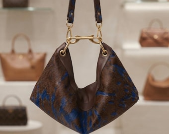 Puede incluir: Un bolso de estilo hobo con una textura de pelo con estampado animal en tonos marrón y azul. El bolso incluye ribetes de cuero marrón liso, herrajes de metal dorado y una correa a juego sujeta con cierres dorados y una barra central.