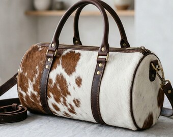 May include: A cylindrical duffel bag crafted from brown and white cowhide with a natural spotted pattern. The bag features dark brown leather handles, matching leather trim, brass-toned metal hardware, and a detachable shoulder strap.