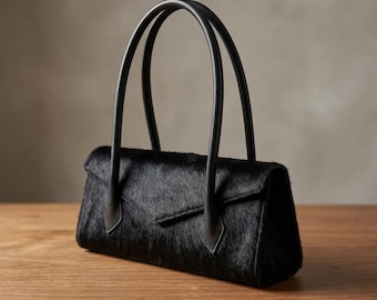May include: A black handbag with a textured hair-on-hide finish, featuring a structured rectangular body, a pointed envelope-style flap closure, and two tall, slender, smooth black leather handles attached to the front and back panels.