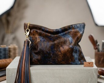 May include: A rectangular clutch bag made of brown and blue cowhide with a textured fur finish. A large leather tassel in matching brown and blue tones hangs from a gold-tone metal clasp on the side. A small rectangular tag reads ARTISAN CO.