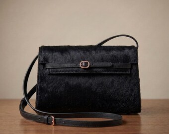 May include: A black hair-on-hide crossbody bag with a rectangular silhouette and a flap closure. The bag features a thin horizontal strap across the front secured by a small rose gold-toned metal buckle and a matching adjustable shoulder strap.