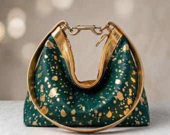 May include: A crescent-shaped handbag featuring a deep green base with a metallic gold splatter pattern. The bag has a smooth gold-toned handle and a central metal clasp closure at the top, connecting the two sides of the curved frame.