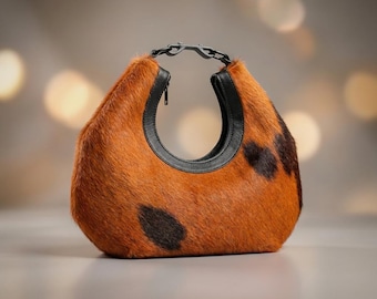 May include: A crescent-shaped handbag crafted from brown and black cowhide with a textured fur finish. The bag features a black leather trim along the circular opening, a black zip closure, and a black plastic link handle at the top.