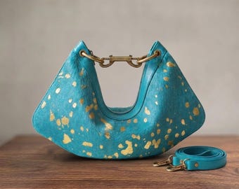 May include: A crescent-shaped handbag crafted from teal-coloured hair-on hide with a metallic gold splatter pattern. The bag features a central brass-toned metal clasp closure and includes a detachable matching teal leather shoulder strap.