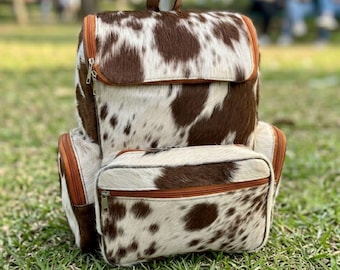May include: A backpack featuring a brown and white cowhide pattern with a textured fur-like finish. The bag includes a large main compartment with a top flap, a smaller front pocket, and two side pockets, all secured with brown zips and metal pulls.