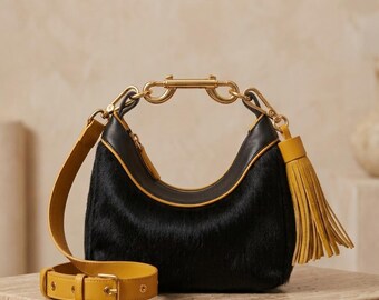 May include: A black hobo bag with a textured hair-on-hide finish, featuring mustard yellow leather trim, a matching adjustable shoulder strap, and a large mustard yellow leather tassel. The bag has gold-tone hardware and a rigid metal handle.