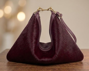 May include: A deep burgundy, triangular-shaped handbag crafted from textured hair-on hide. The bag features a distinctive U-shaped cutout at the top, secured by a gold-toned metal clasp, and includes a matching shoulder strap with an adjustable buckle.