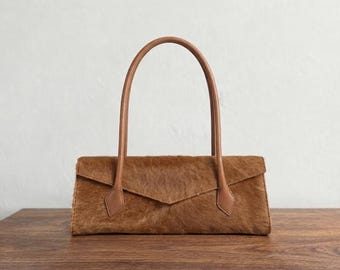 May include: A rectangular handbag crafted from brown textured hair-on hide leather. The bag features a pointed envelope-style flap closure and two thin, rounded leather handles in a matching brown tone, attached to the front panel with stitched tabs.