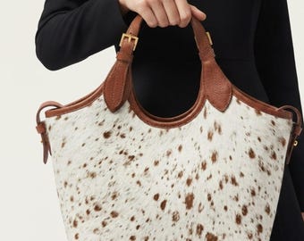 May include: A tote bag featuring a white and brown spotted cowhide pattern with smooth brown leather trim, two leather handles with gold-toned metal buckles, and side leather straps. The bag has a wide opening and a structured, curved silhouette.