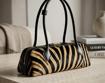 May include: A cylindrical handbag featuring a black and tan zebra stripe pattern with a textured fur-like finish. The bag includes black leather trim, two thin black leather handles, and a gold-toned metal zipper closure along the top edge.