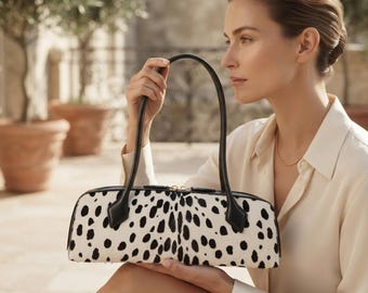May include: A cylindrical handbag featuring a white base with black spots, black leather handles, and a gold-toned zip closure. The bag has a structured, elongated shape with a smooth texture and is held by two thin, black leather straps.