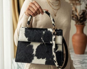 May include: A structured handbag featuring a black and white cow print pattern with a textured hair-on-hide finish. The bag includes a single top handle, a fold-over flap closure, and a detachable shoulder strap with matching cow print detailing.