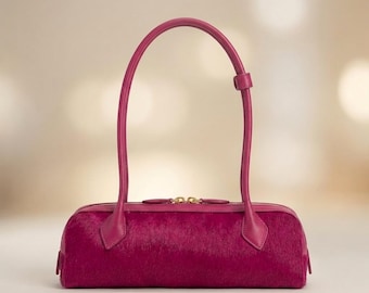May include: A cylindrical handbag in a vibrant magenta hue, featuring a textured hair-on-hide finish. The bag includes a single long, arched handle with a small sliding loop and a top zip closure with gold-toned hardware accents.
