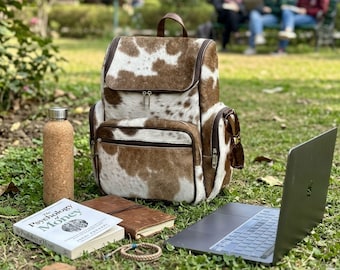 May include: A brown and white cowhide backpack with a top handle and zippered compartments. Beside it are a cork-textured water bottle, a book titled The Psychology of Money, a brown leather notebook, a braided bracelet, and an open laptop.