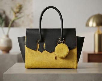 May include: A structured handbag featuring a black leather upper section and a textured yellow hair-on-hide lower body. The bag includes two black top handles and a small, matching circular yellow coin purse attached to the front by a gold ring.