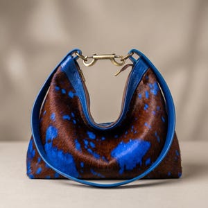 Può includere: Una borsa a spalla in stile hobo realizzata in pelle di vacchetta marrone e blu con pelo. La borsa presenta un cinturino in pelle blu liscia e una chiusura centrale in metallo color ottone sulla parte superiore che unisce i due lati.
