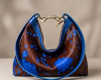May include: A hobo-style handbag crafted from brown and blue cowhide hair-on leather. The bag features a smooth blue leather strap and a central brass-toned metal clasp closure at the top, connecting the two sides of the arched opening.