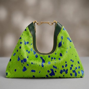 May include: A lime green handbag with a textured hair-on-hide finish and irregular blue paint-like splatters. The bag features a dark green leather trim along the top opening and a gold-toned metal hardware clasp engraved with the text FREDNIC LANCHOS.