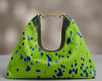 May include: A lime green handbag with a textured hair-on-hide finish and irregular blue paint-like splatters. The bag features a dark green leather trim along the top opening and a gold-toned metal hardware clasp engraved with the text FREDNIC LANCHOS.