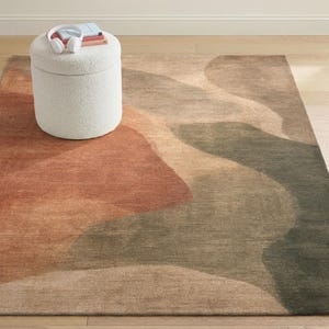 a rug with a colorful abstract design, a white ottoman with a book and headphones on top, and a wooden floor in the background.