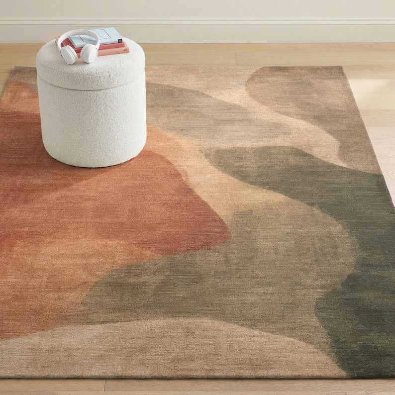 a rug with a colorful abstract design, a white ottoman with a book and headphones on top, and a wooden floor in the background.