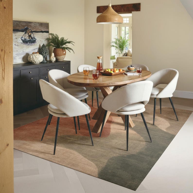 a modern dining room with a wooden table surrounded by white chairs. The table is set with a tray of fruit and drinks, and there are potted plants and a painting on the wall. The room has a neutral color scheme with a colorful rug on the floor.