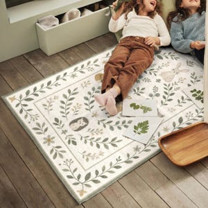A young girl is lying on a rug with a forest-themed design, surrounded by other items on the floor.
