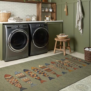 a laundry room with two washing machines and a wooden stool. The room has a green color scheme, and there is a rug with a fish design on the floor.