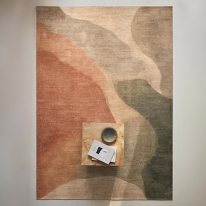 a rug with a gradient design in shades of brown and orange, placed on a wooden surface. There is a small wooden table with a bowl and a book on it, positioned in the center of the rug.