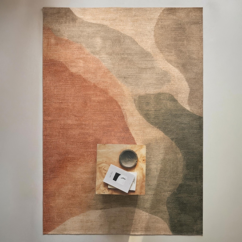 a rug with a gradient design in shades of brown and orange, placed on a wooden surface. There is a small wooden table with a bowl and a book on it, positioned in the center of the rug.