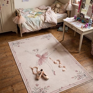 May include: A rectangular area rug featuring a light pink background with a central illustration of a ballerina in a pink tutu. The border is decorated with floral motifs and ballet slippers. A pair of satin ballet shoes rests on the rug surface.