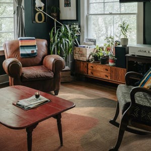 a cozy living room with a brown leather chair, a red coffee table, and a wooden side table with a plant. The room is decorated with various plants and has a window overlooking trees outside.