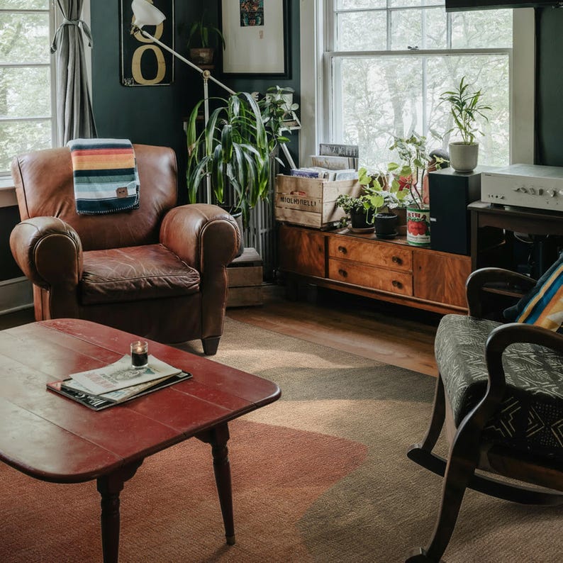 a cozy living room with a brown leather chair, a red coffee table, and a wooden side table with a plant. The room is decorated with various plants and has a window overlooking trees outside.