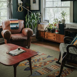 a cozy living room with a brown leather chair, a red coffee table, and a wooden side table with a plant. The room is decorated with various plants, a patterned rug, and a bookshelf.