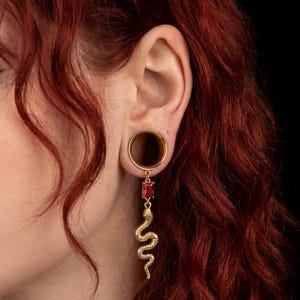 Può includere: Tunnel per l'orecchio dorato con un orecchino pendente. L'orecchino presenta una gemma rettangolare rossa e un ciondolo a forma di serpente dorato dettagliato. Il gioiello è indossato in un lobo allungato, su sfondo scuro.