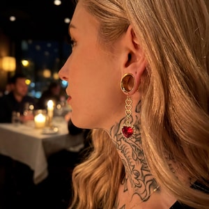 Può includere: Orecchini dorati con un gioiello rosso a forma di cuore e piccoli dettagli a cuore. Gli orecchini sono pendenti e indossati in lobi delle orecchie allungati. L'immagine è scattata in un ristorante poco illuminato.